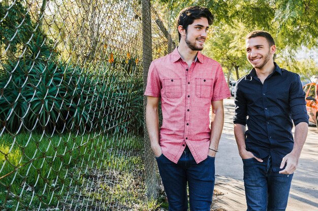 Feliz pareja gay caminando a lo largo de la cerca a la luz del día