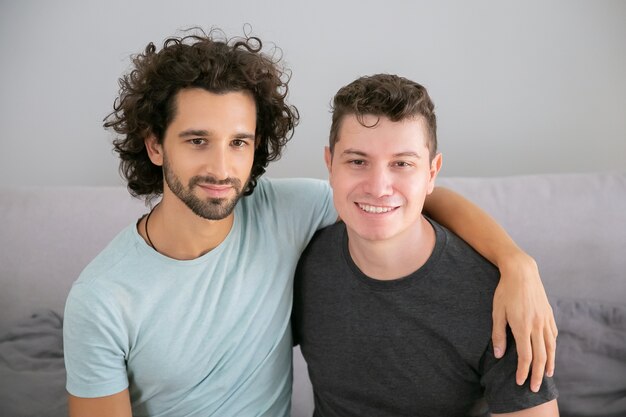 Feliz pareja gay guapo posando en casa, sentados juntos en el sofá y abrazándose. Vista frontal. Concepto de amor y relaciones