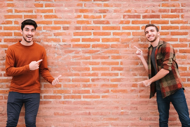 Feliz pareja gay de pie junto a la pared de ladrillo y apuntando con los dedos