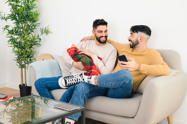 Foto gratuita feliz pareja gay sentada en el sofá mirando el uno al otro con su bebé