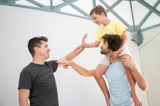 Feliz pareja gay y su hijo alegre divirtiéndose en casa, jugando juegos activos. Niño cabalgando sobre el cuello del hombre y. Concepto de familia y paternidad