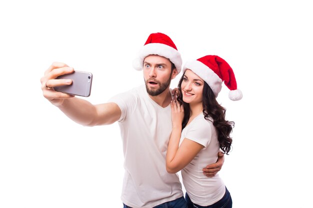 Feliz pareja con gorro de Papá Noel saludando y tomando selfie en smartphone, aislado sobre fondo blanco.