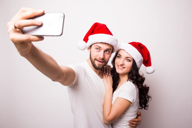 Feliz pareja con gorro de Papá Noel saludando y tomando selfie en smartphone, aislado sobre fondo blanco.