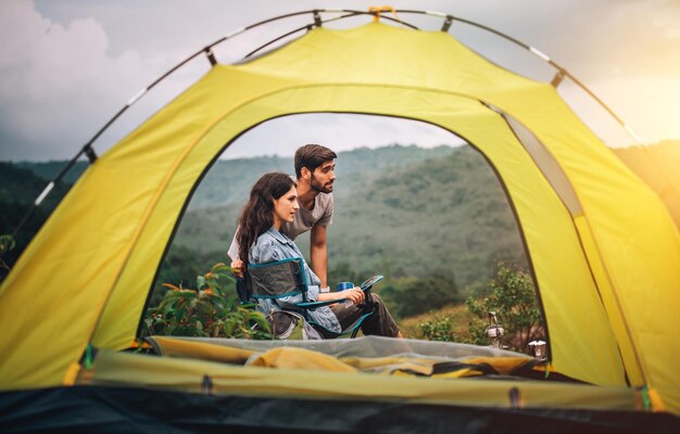 Feliz pareja hombre y mujer sentados en una silla frente a una tienda de campaña en el campamento por la mañana con fondo de montaña