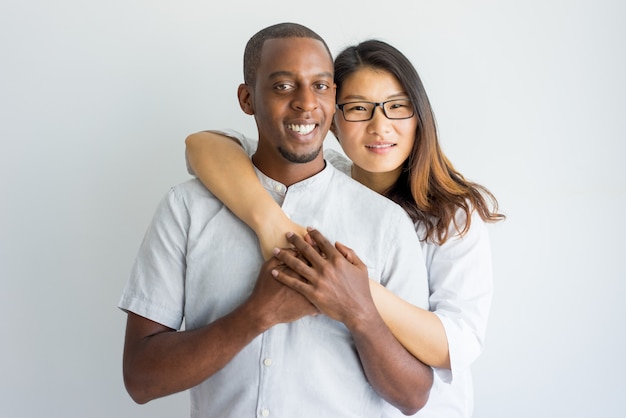 Feliz pareja interracial abrazando y mirando a cámara.