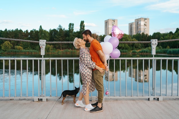 Feliz pareja joven abrazándose y besándose en el puente sosteniendo globos rosas y perro con ciudad en el horizonte