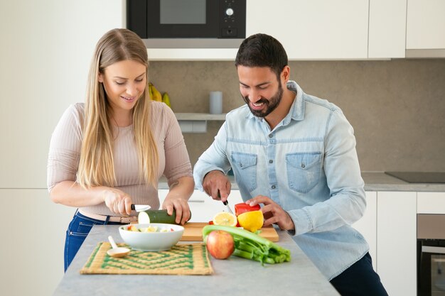 Feliz pareja joven atractiva cocinando la cena juntos, cortando verduras frescas en una tabla de cortar en la cocina, sonriendo y hablando. Concepto de cocina familiar