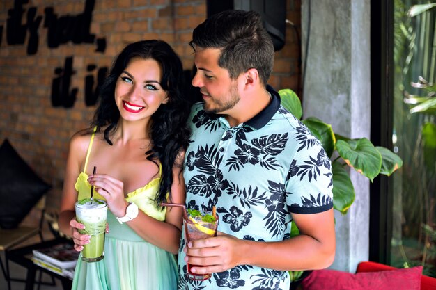 Feliz pareja joven y bonita disfrutando de su sabroso cóctel dulce en el bar tropical, ropa de moda a juego de colores, humor de vacaciones de verano. Cita romántica perfecta.