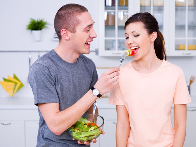 Feliz pareja joven comiendo ensalada en la cocina