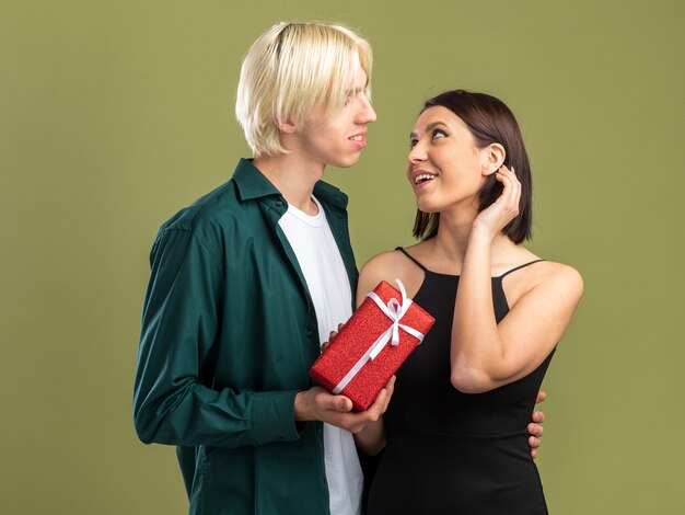 Feliz pareja joven en el día de San Valentín hombre dando paquete de regalo a la mujer que ella toca el cabello ambos mirando el uno al otro aislado en la pared verde oliva