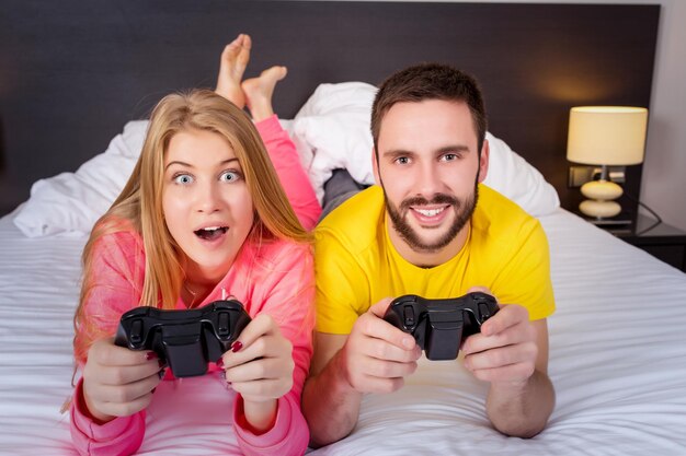 Feliz pareja joven divirtiéndose jugando videojuegos en la cama.