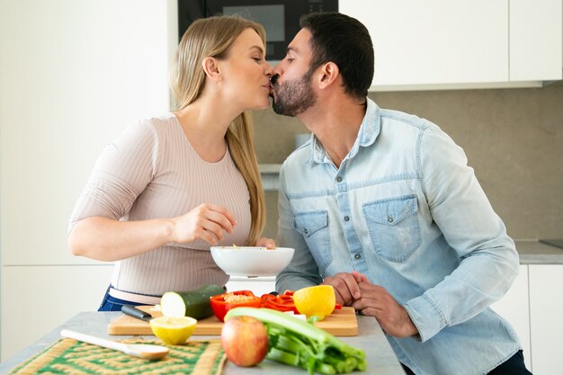 Feliz pareja joven dulce besándose mientras cocina la cena juntos, cortando verduras frescas en una tabla de cortar en la cocina, sonriendo y hablando. Concepto de amor y cocina