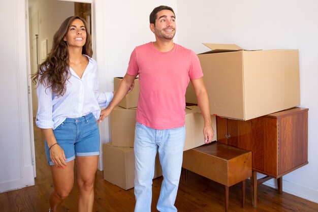 Feliz pareja joven emocionada mirando su nuevo apartamento con cajas de cartón y muebles, sonriendo y hablando