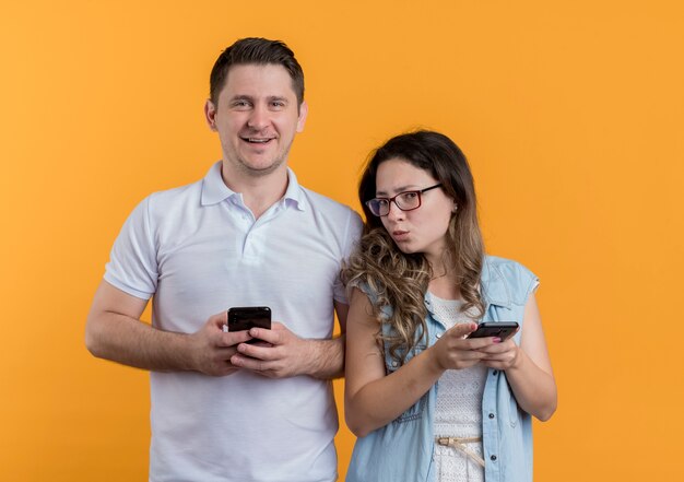 Feliz pareja joven hombre y mujer usando gadgets sonriendo de pie sobre la pared naranja