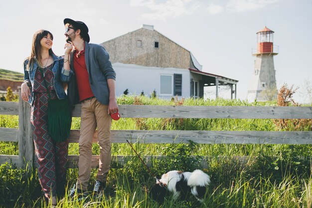 Feliz pareja joven inconformista con estilo en el amor caminando con el perro en el campo, moda boho de estilo veraniego