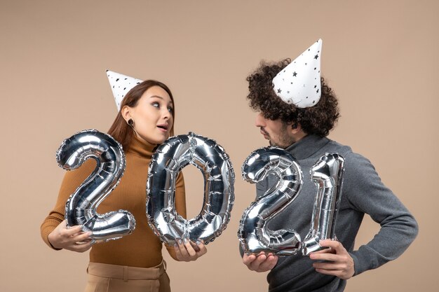 Feliz pareja joven mirándose usar sombrero de año nuevo posa para la cámara Chica mostrando y chico con y en gris
