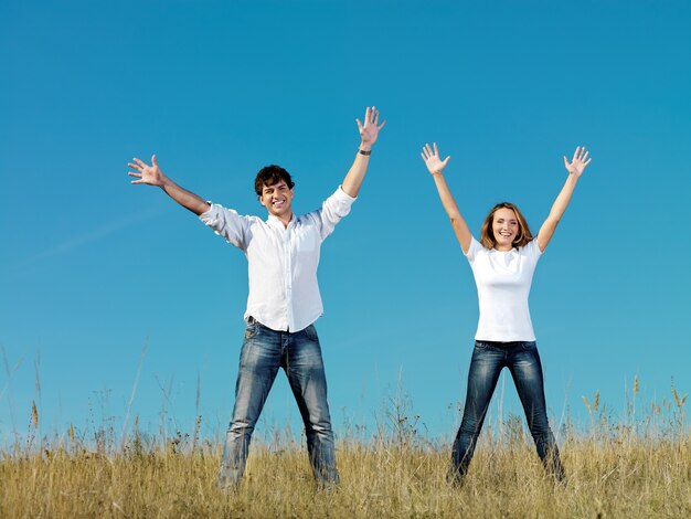 Feliz pareja joven de pie juntos en la pradera de verano con las manos arriba