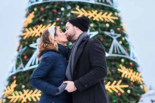 Feliz pareja joven con ropa abrigada parada cerca de un árbol de Navidad de la ciudad, disfrutando pasar tiempo juntos