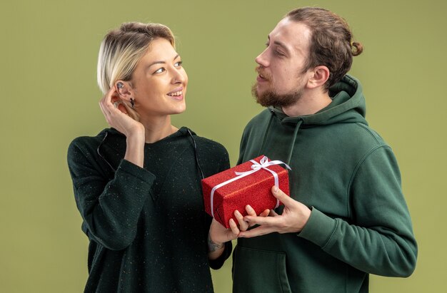 Feliz pareja joven en ropa casual hombre con presente para su encantadora novia sonriente celebrando el día de San Valentín de pie sobre la pared verde