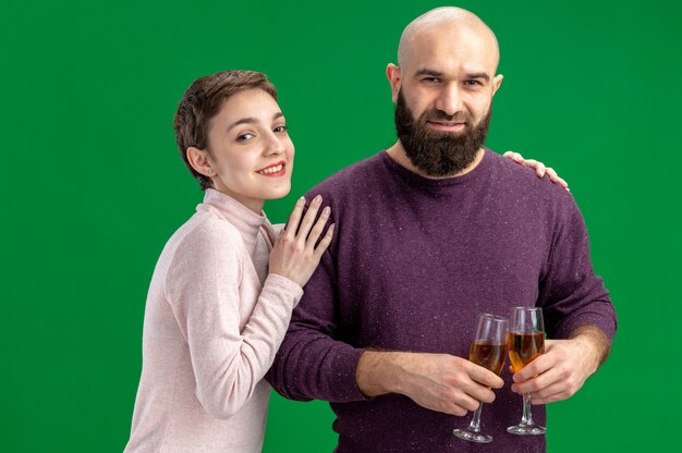 Feliz pareja joven en ropa casual mujer con pelo corto y hombre barbudo con copas de champán felices enamorados juntos celebrando el día de San Valentín de pie sobre la pared verde