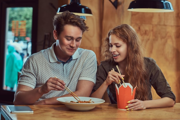 Feliz pareja joven con ropa informal comiendo fideos picantes en un restaurante asiático.