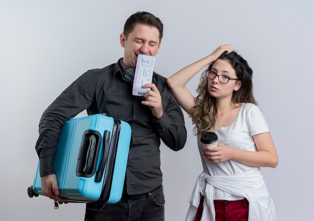 Feliz pareja joven de turistas hombre y mujer sosteniendo maleta y billetes de avión de pie sobre la pared blanca