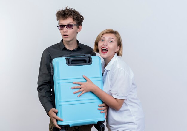 Feliz pareja joven de turistas hombre y mujer sosteniendo la maleta mirando sorprendido y asombrado de pie sobre la pared blanca