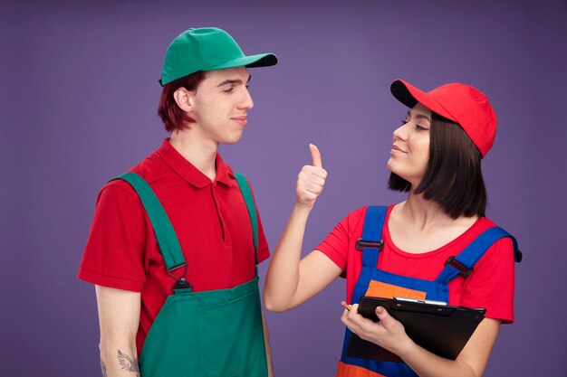 Feliz pareja joven en uniforme de trabajador de la construcción y gorra mirando el uno al otro niña sosteniendo lápiz y portapapeles mostrando el pulgar hacia arriba aislado en la pared púrpura