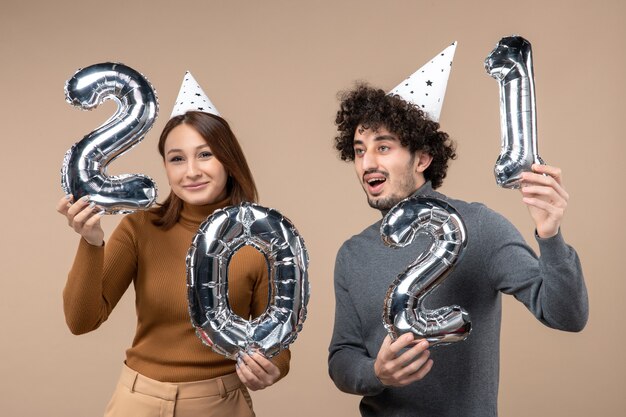 Feliz pareja joven usa sombrero de año nuevo posa para la cámara Chica tomando y chico con y mirando en diferentes direcciones en gris