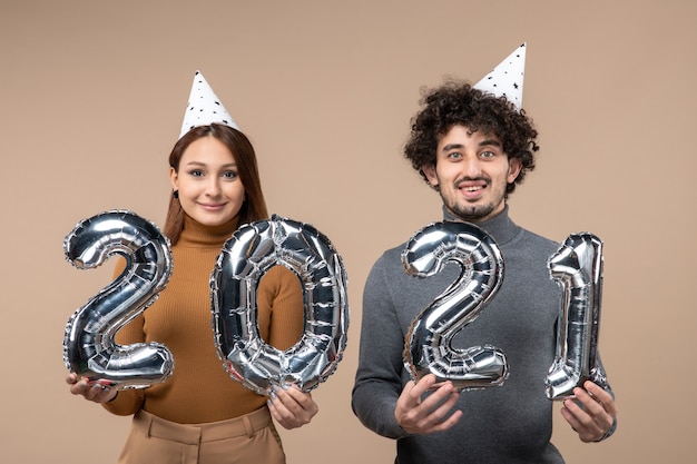 Feliz pareja joven usa sombrero de año nuevo posando con en sus manos en gris