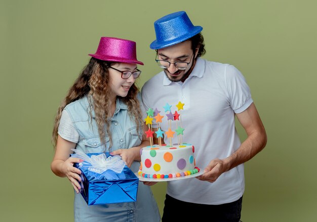 Feliz pareja joven vestida con sombrero rosa y azul chica sosteniendo una caja de regalo y chico sosteniendo y mirando con chica en la torta de cumpleaños