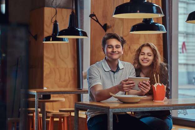 Feliz pareja de jóvenes estudiantes usando una tableta digital durante el almuerzo en un restaurante asiático.