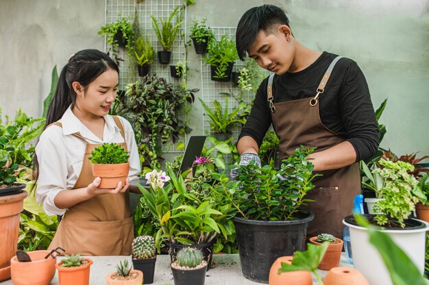 Feliz pareja de jóvenes jardineros asiáticos vistiendo delantal utiliza equipo de jardín y computadora portátil para cuidar