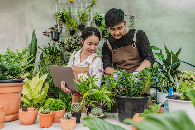 Feliz pareja de jóvenes jardineros asiáticos vistiendo delantal utiliza equipo de jardín y computadora portátil para cuidar