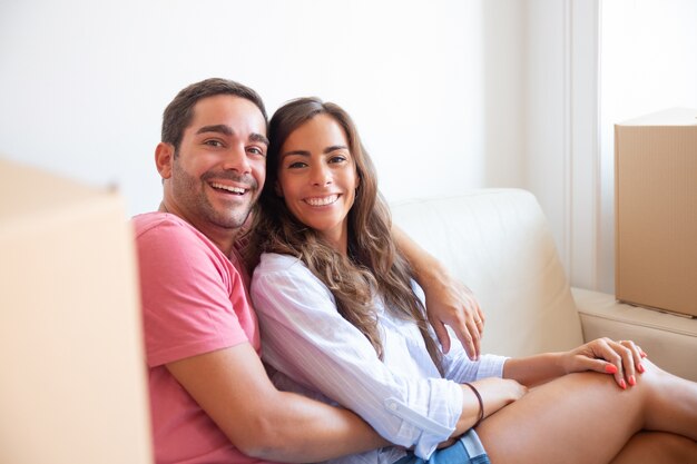 Feliz pareja latina sentada en un sofá entre cajas de cartón en la nueva casa, mirando a la cámara, sonriendo, riendo