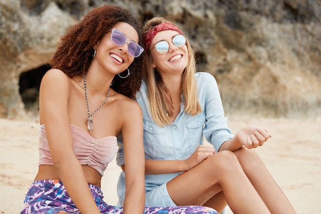 Feliz pareja de lesbianas disfruta del tiempo de recreación en el desierto con expresiones alegres, usan gafas de sol, se sientan contra el acantilado. Jóvenes feministas encantadoras disfruta de la unión en un tranquilo lugar turístico