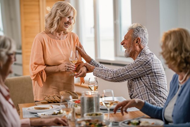 Feliz pareja madura bebiendo vino y divirtiéndose durante el almuerzo con sus amigos en la mesa de comedor