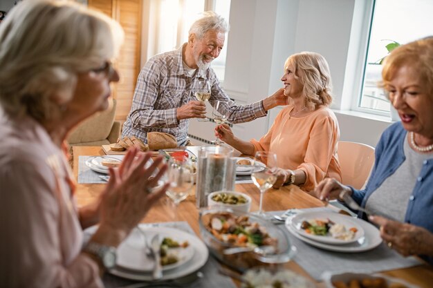 Feliz pareja madura brindando con copas de vino y hablando durante el almuerzo con sus amigos en casa