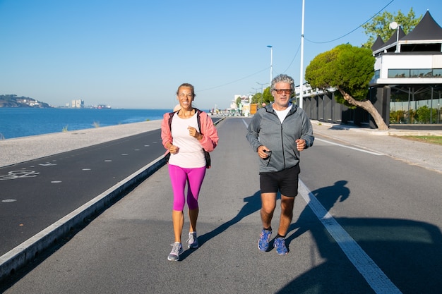 Feliz pareja madura para correr a lo largo de la orilla del río. Hombre y mujer de pelo gris con ropa deportiva, corriendo afuera. Concepto de actividad y edad