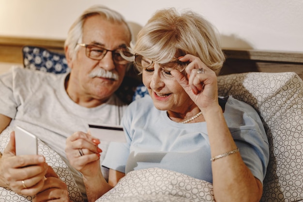 Feliz pareja madura demandando teléfono inteligente y tarjeta de crédito mientras está acostado en la cama El foco está en la mujer leyendo el número de tarjeta de crédito