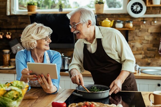 Feliz pareja madura preparando comida y comunicándose mientras usa una tableta digital en la cocina