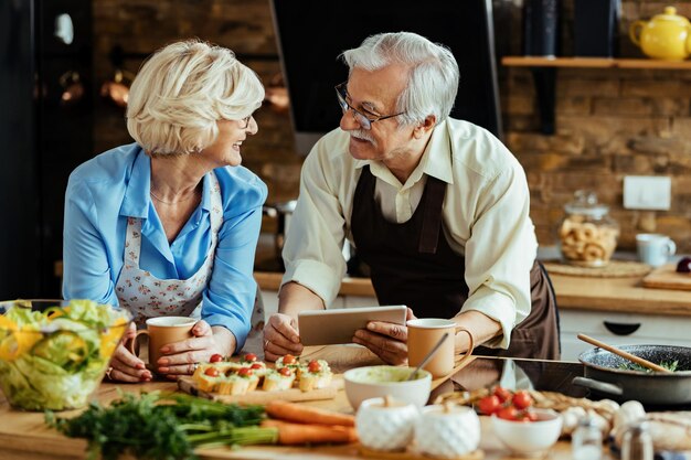 Feliz pareja madura usando tableta digital y hablando mientras prepara el almuerzo en la cocina.