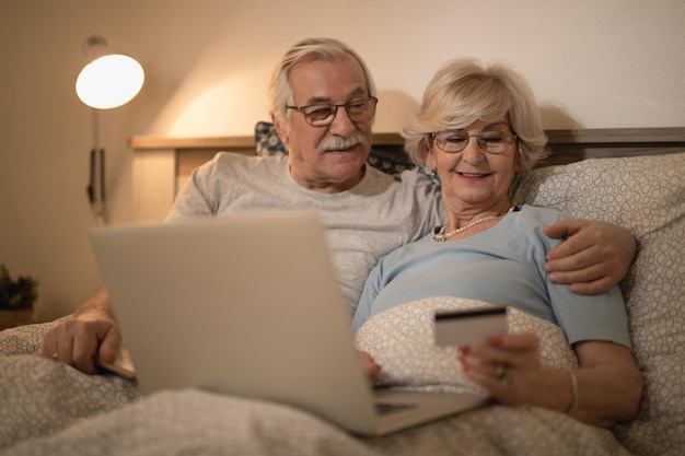 Feliz pareja mayor comprando en Internet con laptop y tarjeta de crédito mientras se acuesta en la cama por la noche