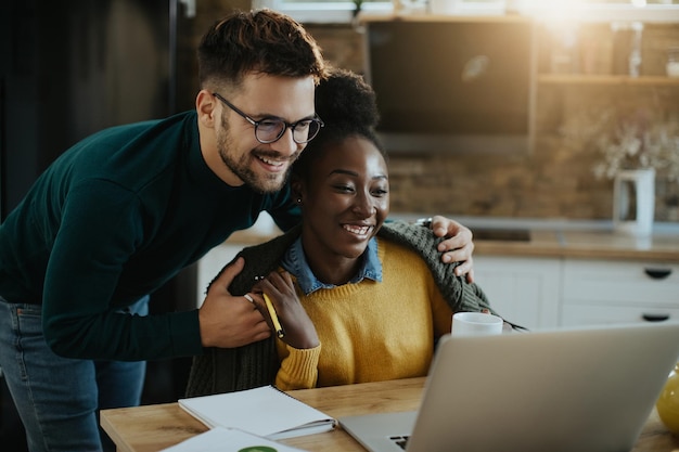 Feliz pareja mixta leyendo un correo electrónico en una laptop en casa