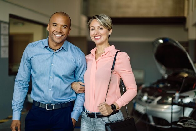 Feliz pareja mixta en el taller de reparación de automóviles mirando a la cámara