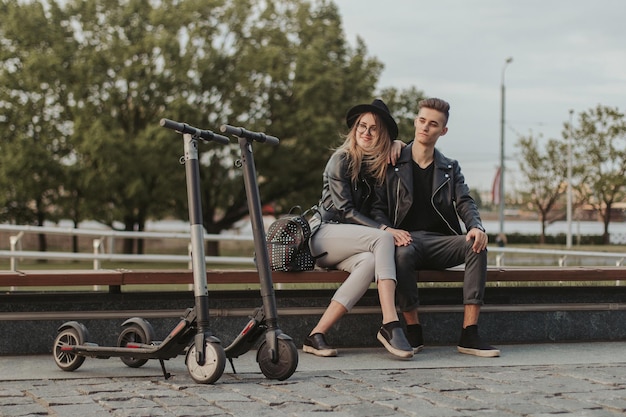 Una feliz pareja de moda se está relajando en el banco del parque de la ciudad con sus scooters.