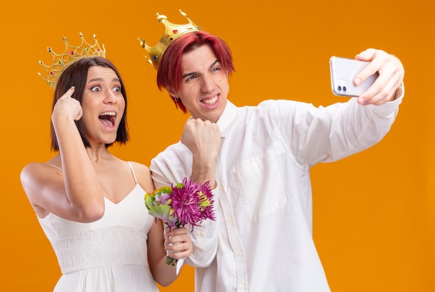 Feliz pareja de novios con ramo de flores en vestido de novia con coronas de oro sonriendo alegremente haciendo selfie con smartphone parado sobre pared naranja