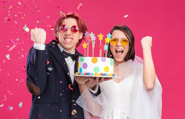 Feliz pareja de novios en vestido de novia con gafas sosteniendo pastel de boda apretando los puños feliz y emocionado sonriendo alegremente de pie en rosa