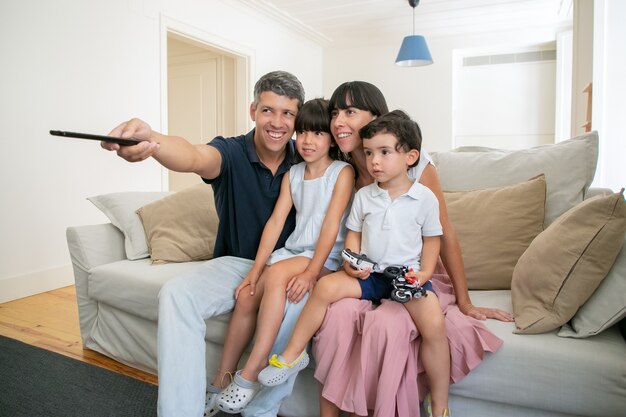 Feliz pareja de padres con dos hijos viendo la televisión, sentado en el sofá en la sala de estar y usando el control remoto.