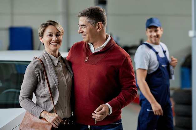 Feliz pareja de pie abrazada en el taller de reparación de automóviles mientras el mecánico de automóviles está en el fondo